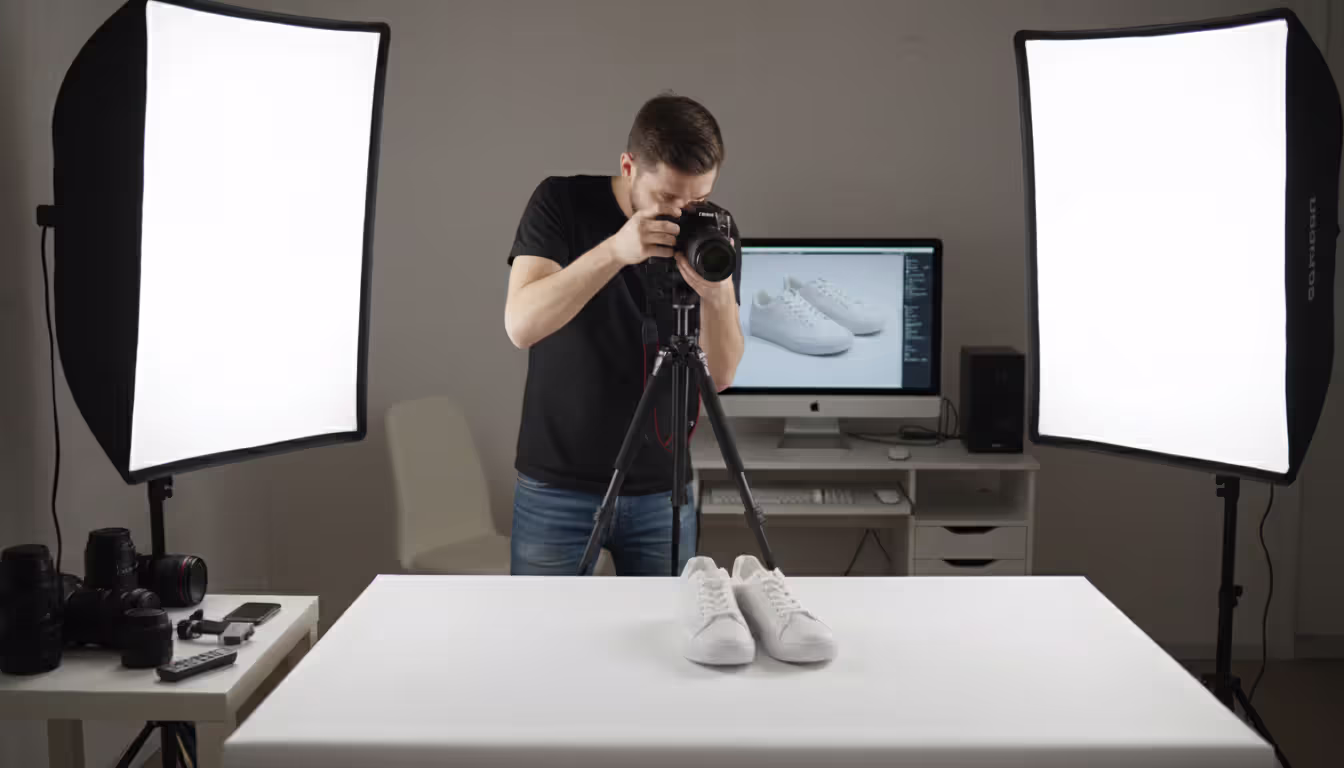 Young photographer shooting product photos of white sneakers on light table with two softbox lights and camera on tripod in home studio
