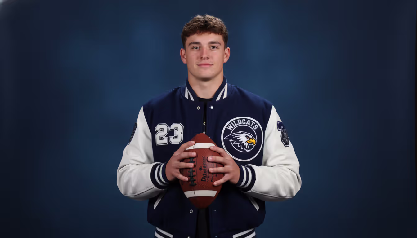 High school senior boy posing in a photo studio wearing a varsity letter jacket and holding a football against a dark blue backdrop