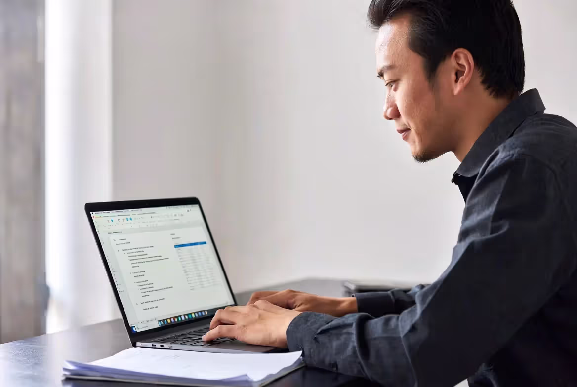 A focused young man sitting at a desk, typing on his laptop while composing a professional document